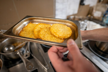 A close-up of hand holding a tray with freshly cooked golden-brown potato pancakes. The background features busy kitchen setting with various cooking utensils visible. Ideal for food-related content.