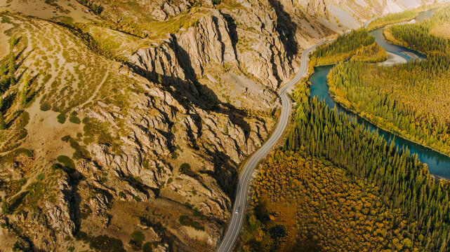 Aerial view of a scenic highway winding through a dramatic mountain valley, following the curves of a turquoise river, amidst colorful autumn foliage