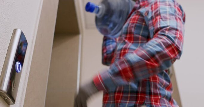Man with water bottle on shoulder calls lift using button. Delivery service courier fulfills order carrying container to apartment low angle shot
