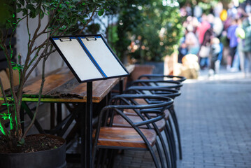 outdoor restaurant setting with wooden tables, metal chairs, and a blank menu stand in a vibrant urban street, surrounded by greenery and blurred figures of people in the background