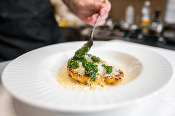 A chef garnishing a beautifully plated dish with fresh green sauce and grated cheese, showcasing culinary expertise and attention to detail in a modern kitchen setting.