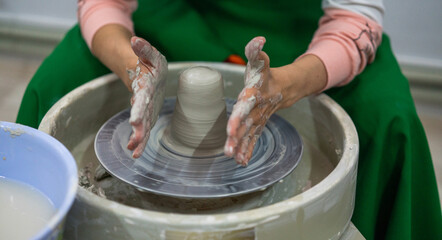 A person wearing a green apron with hands covered in clay is shaping a piece of pottery on a spinning wheel. Fully engaged in the creative process with a focus on the clay piece.