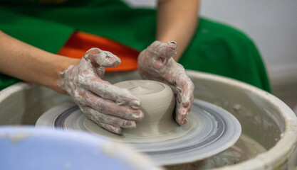 A person wearing a green apron with hands covered in clay is shaping a piece of pottery on a spinning wheel. Fully engaged in the creative process with a focus on the clay piece.