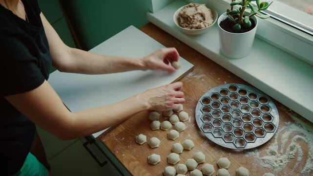 Woman arranging freshly made dumplings on board for preparation. Female organizing dumplings after shaping them using metal mold. Lady placing dumplings in rows on floured board beside kitchen window