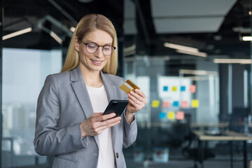 Businesswoman in business suit standing by window inside office with phone in hands and bank credit...