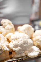 Fresh, raw cauliflower heads are displayed on a tray, showcasing their white, textured surface. The background is softly blurred, emphasizing the vegetables in the foreground