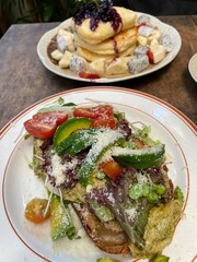 A colorful and appetizing lunch spread featuring a fresh vegetable topped toast with creamy pesto and parmesan, paired with a stack of fluffy pancakes garnished with fruit and berry sauce