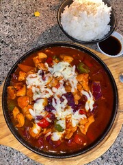 A sizzling hot plate of Korean-style spicy chicken stew topped with melted cheese, served with a bowl of steamed white rice and a side of soy sauce