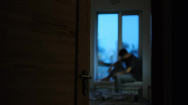 A man sits on a windowsill in a dark room, viewed through an open door, expressing emotional isolation and sadness.