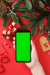 Festive Christmas smartphone mockup with green screen in hand on red background with fir branches, pinecones, tags, ribbon and small gifts