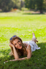 Cheerful woman in white dress lying on the grass, smiling with sunglasses on her head, enjoying a sunny summer day in the park.