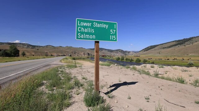 A road sign on Idaho State Highway 75 at Stanley&rsquo;s entrance shows distances to towns like Challis and Salmon. The Salmon River flows by, cattle graze, and a truck with a trailer passes.
