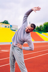Cheerful caucasian male in grey activewear bending and doing warming exercising on stadium for losing weight and slimming, positive strong sports man prepare for cardio training enjoying workout