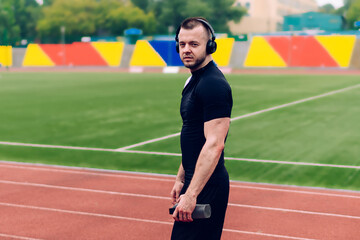 Obraz premium Handsome caucasian male in trendy wear and headphones walking on stadium listening favorite songs, portrait of hipster guy prepare for cardio workout enjoying online radio in sound accessory