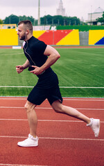 Young handsome male in earphones running on stadium dressed in active wear reaching sport goals, strong man athlete keeping fit having morning cardio training with favorite music in headphones