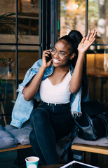 Smiling african american woman in casual wear sitting in cafe interior talking on mobile phone and waving to somebody, cheerful dark skinned female satisfied with positive smartphone conversation