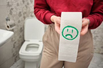 Young woman holding toilet paper roll with sad smiley face drawn on it near toilet in bathroom.