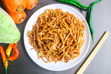 Top view of spicy stir-fried noodles with vegetables like cabbage and carrots and chili in a white plate on a gray background. Indonesian food fried noodles