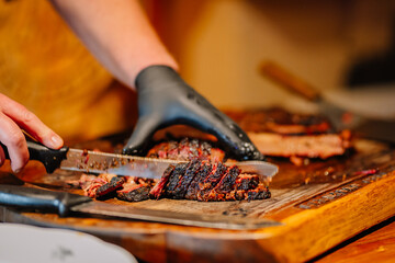 A close-up of a person slicing smoked, charred meat with a knife on a wooden cutting board, wearing black gloves for handling food.