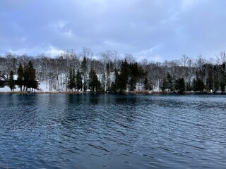 Beautiful lake scene at Green Lakes, Syracuse New York.  Treelined lake with snow covering the ground and calm waters.  Cloudy sky. No people. Copy Space