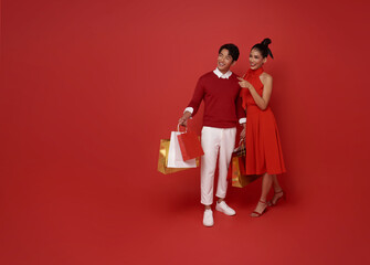 Happy asian couple in red casual attire holding shopping bag and shopping in chinese new year day isolated on red background.
