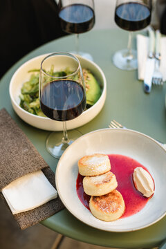 Quark pancakes (Syrniki) with strawberry jam, an avocado salad and three glasses of red wine on a table at an outdoor cafe in summer