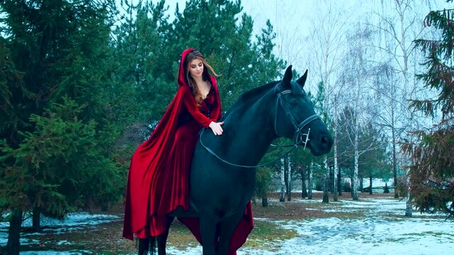 medieval fantasy woman rider sits astride on horseback, stroking black horse pet animal mare. Fairy elf Girl princess Enjoy walking winter nature snow forest green trees. Long red cape Hood on head