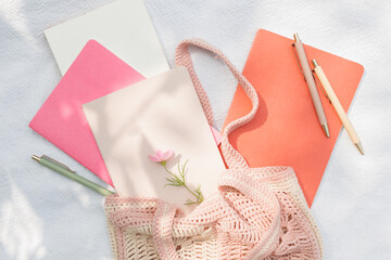 Overhead view of assorted notebooks, pens and a pink flower in a pink knitted bag on white fabric