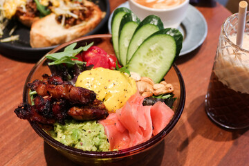 close up of roasted chicken and cashew nut salad, with pickled ginger, cucumber, candied tomato, green vegetables, smashed avocado and turmeric tahini in a transparent bowl