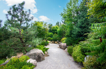Sun shines down on a stone and gravel path through a Japanese-inspired garden with various plants and foliage on a sunny summer day in Grand Rapids, Michigan.
