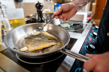 A close-up of a chef's hand spooning sauce over two fish fillets cooking in a pan. The kitchen setting features an elegant, modern look with various cooking tools in the background.