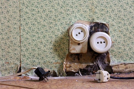 A Pair Of Vintage Electrical Outlets With Fraying Plugs, Set Against Old Floral Wallpaper. The Image Illustrates Aging Infrastructure And Potential Electrical Safety Hazards.
