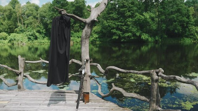 The knight's sword on the pier of the river. Steel sword in a sheath, medieval weapon and cloak on a tree branch on the pier, dock for ships. Equipment of a warrior monk in ancient times.