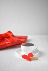 A white cup of coffee, a red paper heart next to it, and red gift boxes in the background. The concept of Valentine's day.