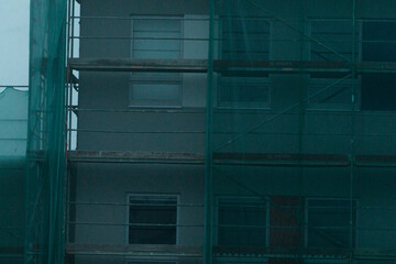 Building is under construction and features green protective netting draped across its facade for safety and protection purposes. Foggy urban background, dark winter day.