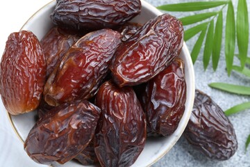 Many tasty dried dates in bowl and leaves on gray textured table, flat lay