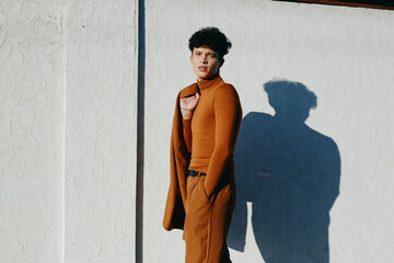 Young man in a stylish, form fitting brown outfit leaning against a textured white wall, showcasing...