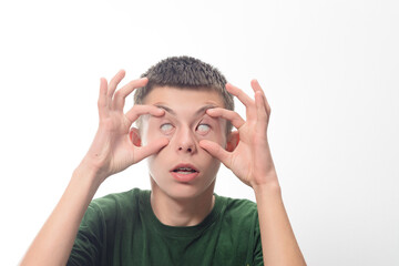 A young man playfully pulls at the corners of his eyes, creating a comical expression. The image is set against a plain white backdrop, emphasizing the facial features.