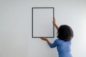 Brunette Female Hanging Empty Picture In Frame On White Wall Decorating House Standing Indoors. Modern Art And Home Interior Decor. Selective Focus On Blank Poster. Mockup. High quality photo