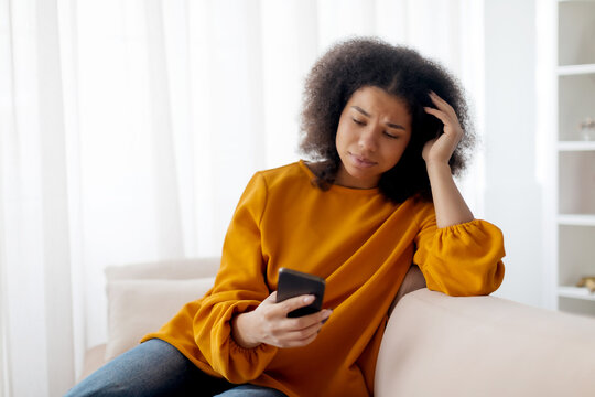 Worried african lady looking at smartphone screen, sitting on couch at home, concerned african american woman waiting for important call, reading unpleasant sms, spending time with phone in living