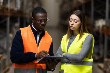 Two warehouse workers wearing safety vests discuss logistics while reviewing notes on a clipboard, standing in a large storage facility. The serious expressions 