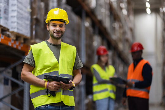 Warehouse worker smiling while holding scanner in a busy storage facility - Powered by Adobe