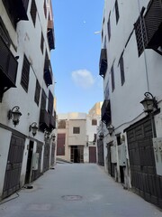 Street of old Jeddah. On both sides there are facades of white old buildings with brown wooden balconies. Antique iron lanterns hang on the facades of buildings. Blue sky with clouds