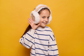 A girl with long hair wears fluffy earmuffs while smiling and playfully raising her hand to her ear. The bright yellow background adds warmth to the winter vibe