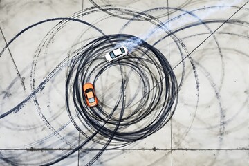 Aerial view of two cars drifting on concrete, leaving circular tire marks, with smoke trails visible.