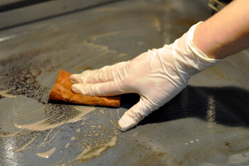 A woman's hand in a latex protective glove washes a dirty oven with a sponge. The sponge is torn. Heavy pollution.