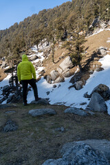 A traveler in a neon green jacket stands on a rocky patch of grass, gazing at a snow-covered...