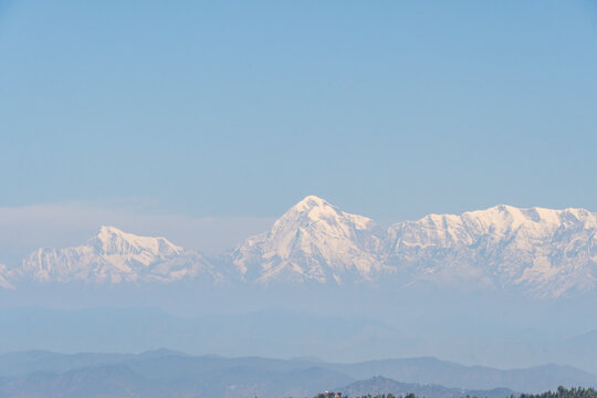 A stunning view of a snow-capped mountain range under a clear blue sky. The peaks stand majestically with layers of misty hills in the foreground. A serene and tranquil landscape in Mukteshwar, India.