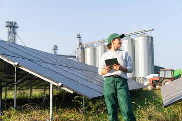 Female farmer with digital tablet on a modern farm using solar panels. Agricultural silos in the...