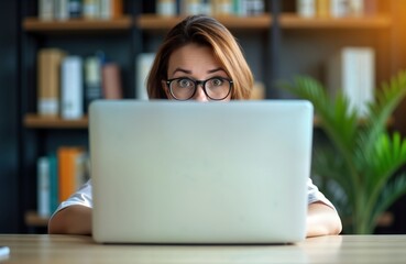 Woman hides behind laptop looking at colleagues. Confident business lady peeks over laptop at workplace. Focused expression. Modern workspace with wooden table, bookshelf in background. Possible
